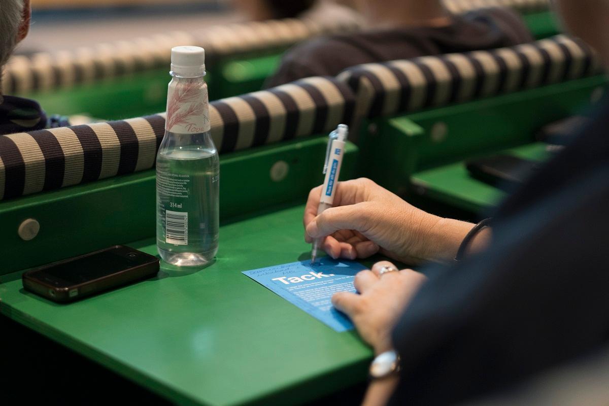 Kvinnas hand syns skrivandes på papper under kongress. Vattenflaska och mobiltelefon.